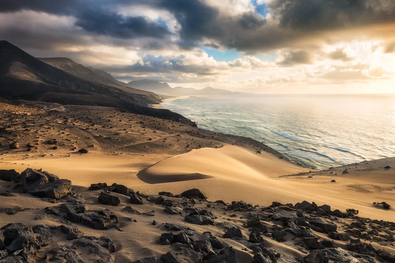 Dark rocks, golden dunes, Mountains, deep clouds, the sea and golden light at Jandia National Park