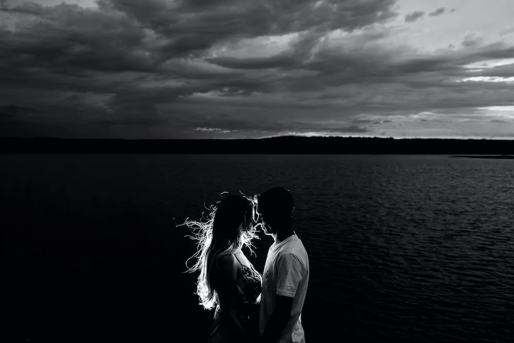 Backlit couple in black and white photograph sharing an intimate moment at night on the water