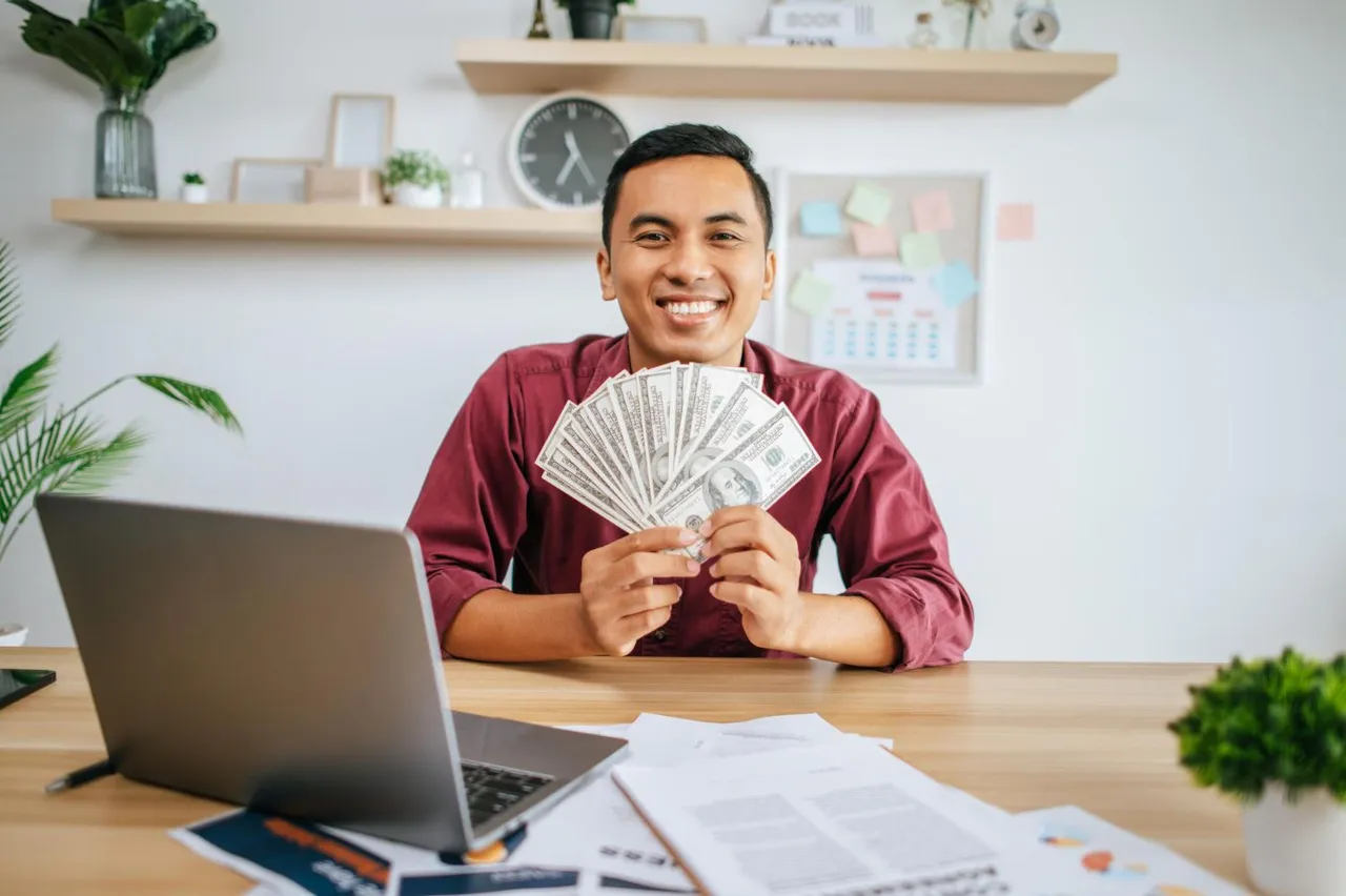 man-working-office-holding-money-with-laptop-documents-desk.jpg