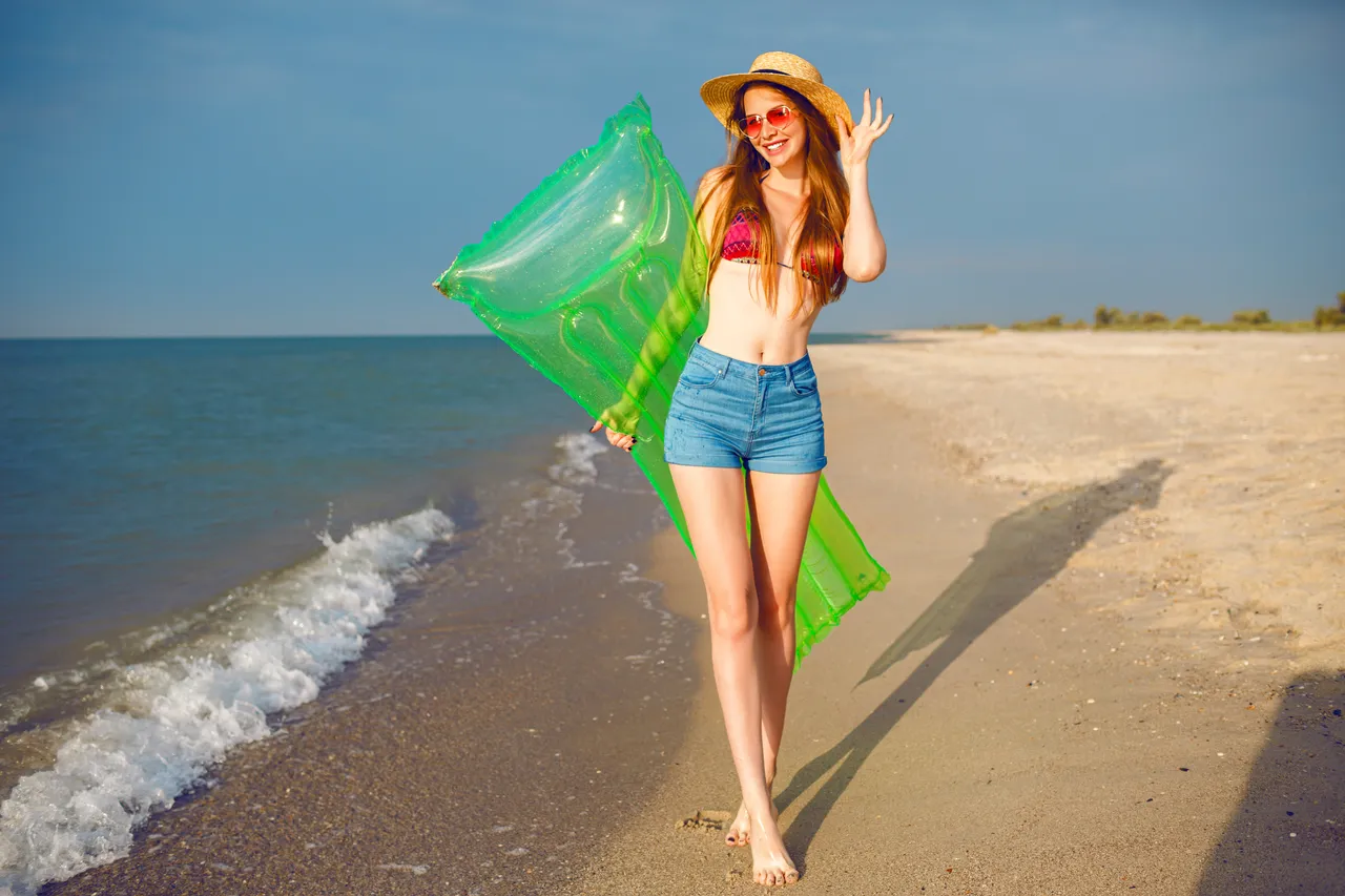 happy_pretty_woman_having_fun_beach_wearing_stylish_beachwear_bikini_hat_denim_shorts_long_legs_slim_fit_body_holding_air_mattress_walking_near_ocean.jpg