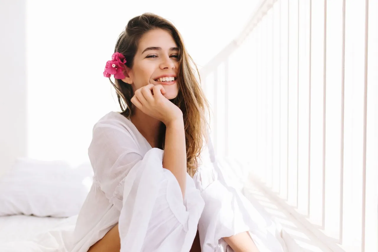 romantic-girl-with-sly-smile-in-vintage-blouse-sitting-on-bed-and-touching-her-chin-with-hand-portrait-of-dreamy-cute-young-woman-with-flower-in-hairstyle-resting-in-bedroom-in-morning.jpg